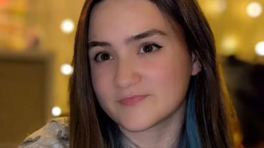 Seren Jaye is pictured with long, straight brown hair with a streak of blue stands indoors against a warm, softly lit background featuring circular light spots. She is wearing a light-coloured top with a subtle pattern. 