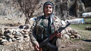 Female Iranian Kurdish fighter dressed in headscarf and grey uniform and holding an automatic weapon stands in front of a pile of stones and some tents in the distance.