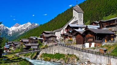 A general view of a row of buildings in Blatten, on an elevated road overlooking a stream. There are forest-covered mountains behind. The sky is bright blue and there are snow-topped mountains in the background.