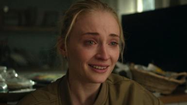 Picture of Sophie Turner looking upset, with tears in her eyes and her hair pulled back. She is wearing a khaki bomber jacket that is slightly unzipped. Messy blurred household items appear in the background.
