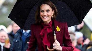 Catherine wearing burgundy with a yellow daffodil pinned to her blazer, holding a black umbrella, as people take pictures in the background