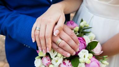 A stock image showing the hands of a bride and groom, resting on a bouquet of flowers and wearing wedding rings,