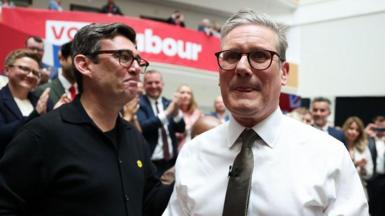 A smiling Andy Burnham looks at Sir Keir Starmer, who is licking his lips. They are the launch of Labour's general election manifesto