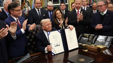 US President Donald Trump signs document to end shutdown, holding it up at his desk which a crowd of people around him applaud.