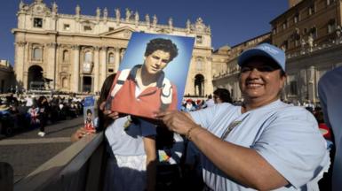 A woman in a crowd at the Vatican, wearing a white T-shirt and a pale blue baseball cap holds up a portrait picture of curly-haired Carlo Acutis, wearing a red jumper with blue collar, with the white handles of a backpack showing