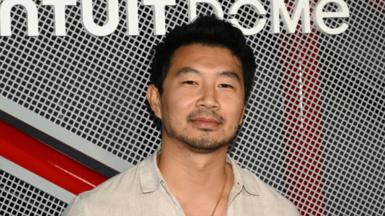 Actor Simu Liu smiles at the camera. He is wearing a beige shirt and a gold necklace.