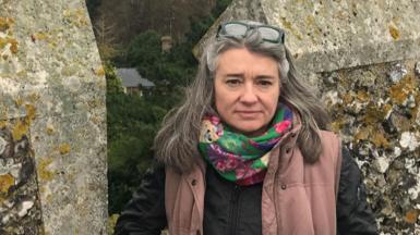 Monique Bonney in front of a stone wall. Behind her is a view of north Kent, including fields, a house and in the distance a town.