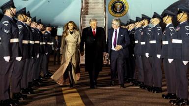 Donald Trump holds hands with First Lady Melania Trump as they walk through a column of men in military dress alongside Viscount Henry Hood. Air Force One is behind them with its steps extended.