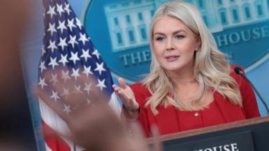 Karoline Leavitt taking questions from the press in the White House. She is wearing a red suit.