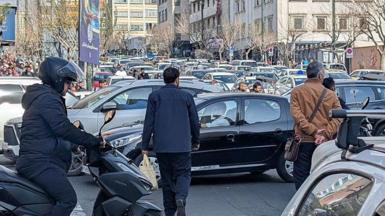 Iranians try to clear a street amid heavy traffic in Tehran, Iran, on February 28, 2026. One man on a motor scooter and two others on foot stand around with rows of stationary cars in the background on a city street.