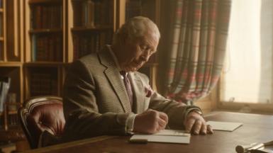 King Charles III writes a letter from a desk in front of a window in Buckingham Palace. He is wearing a blazer and a purple tie. 