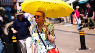 A women holds a yellow umbrella to shade her from the sun. She walks along a high street in the UK wearing a summer dress and carrying a red handbag. A man behind her is wearing a hat and a dark top. 