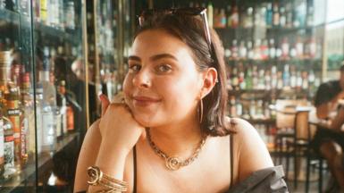 A young woman in a bar with lots of bottles of drink in cabinets behind her. You can see the top half of her body. She is wearing a black top and resting her chin on her hand. She has sunglasses pinning back her hair and has golden earings, necklace and bracelet. 