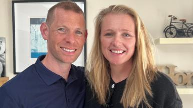 Man smiling at camera wearing blue shirt. Women smiling at camera wearing a black jumper. There are shelves in the background and a cream wall uwith a picture frame. 