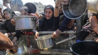 Several people holding pans to fill with food at an aid distribution site - with one woman in the middle wearing all black visibly upset and crying