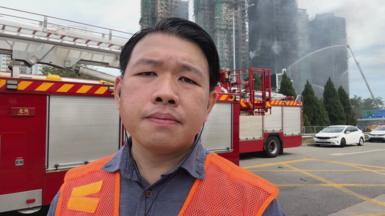 A man in an orange vest stands in front of a red fire engine, with three tower blocks seen smoking in the distance.