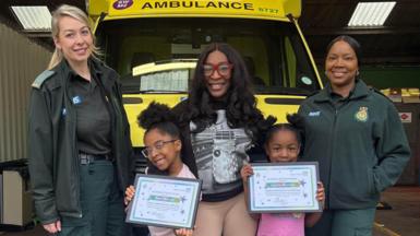 Two smiling little girls hold framed certificates of commendation. Their mother Carole is standing smiling behind them. They are in front of a yellow London ambulance and paramedic Charlotte Aisbitt and call handler Gen Hinds are dressed in their uniform of dark green top, jacket and trousers and are standing to their left and right smiling.