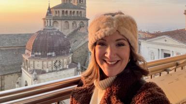 Monica Stott in front of a cathedral in Bergamo, Italy. She is smiling at the camera and wearing a cream hat and brown coat.