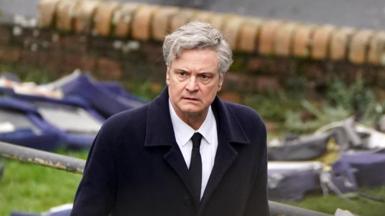 Colin Firth walking with a concerned expression. Wreckage can be seen in the background, and he is wearing a black coat and tie, with a white shirt.