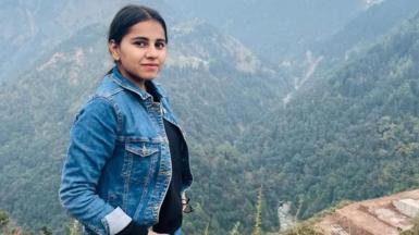 Harshita Brella, who has tied-back black hair and is wearing a denim jacket and a black top. She is posing for a photo in a high spot with tree-covered mountains behind her.