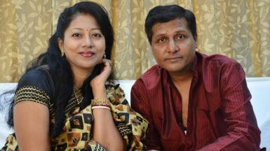 Sougat Mukherjee and his wife, Sapna. They both have dark hair - hers is long - and they are both wearing traditional Indian dress and looking at the camera. 