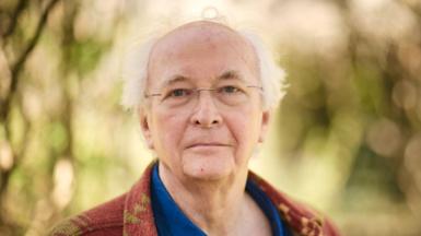 Philip Pullman with glasses in a brown & beige cardigan and blue shirt against trees and leaves background