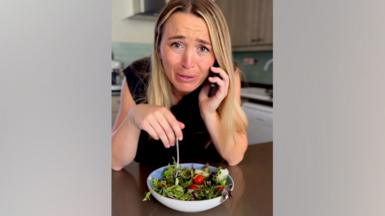 Person sitting at a kitchen table holding a mobile phone to one ear while eating salad