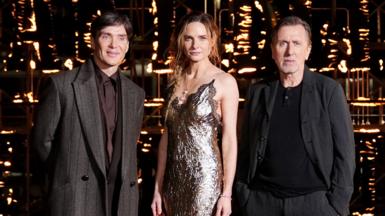 A man with dark brown hair in a tweed coat and purple shirt, a woman with long blonde hair in a silver dress and a man with dark hair a black top and dark jacket at a film premiere