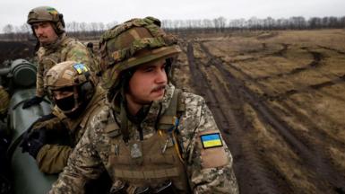 Ukrainian soldiers in full battle dress sit on a cannon in a muddy field.  