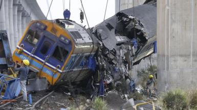 Rescuers work the wreckage of a passenger train after a construction crane collapsed onto it. The train is partially suspended, with parts of it completely crushed