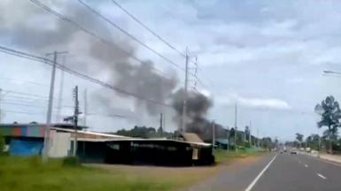 Smoke rises from a structure, amid the clashes between Thailand and Cambodia, in Kantharalak district, Sisaket province, Thailand. Black smoke rises up into the blue sky. Other smaller structures can be seen in the foreground of the picture, which is a still from a video. A tarmac road runs alongside one side of the picture.