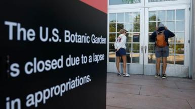 Two visitors in shorts and sweatshirts look through the door windows of the closed US Botanical Gardens during the federal government shut down. In the foreground is a black sign with white lettering reading: 'The US Botanic Garden is closed due to a lapse in appropriations'. 