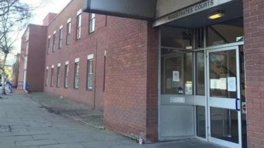A red-bricked double-storey building with a large glass door. Above the door is a sign that reads: "Magistrates' Courts". There are paving slabs leading up to the entrance. 