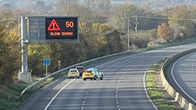 A view of an empty M5 motorway, with a "50mph slow down" matrix sign active. There are two police cars parked to one side of the road with a third dark coloured car in front of them.