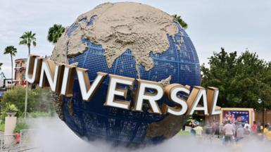 A large model of a globe stands within a theme park. Around the Equator is the logo "UNIVERSAL" in capital letters. There are some people in shot, along with some palm trees.
