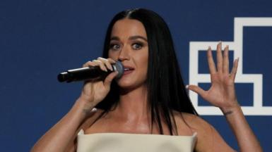 Katy Perry holding microphone to her mouth in right hand with left hand raised and open