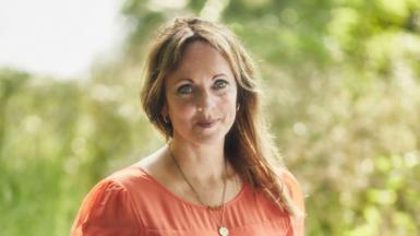 Jenny Evans smiles at the camera. She is wearing a coral pink dress and a necklace. Her blonde hair is pulled to the side and is resting on her shoulder. Behind her is a blurred background of a garden.