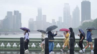 People with umbrellas walk over a bridge in a rain storm with skyscrapers and a river and another bridge in the background