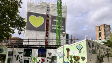 A partially demolished Grenfell tower with a wall surrounding the area which is covered in tributes and drawings. The building is covered in a wrap which features a green heart.