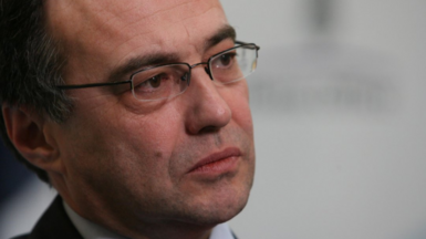 Phil Woolas, wearing a suit and glasses, looks thoughtful as his photo is taken by a professional photographer. He is photographed partly side on looking to the right of the frame.