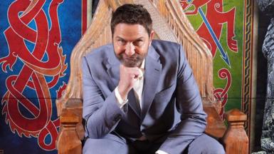 A man in a light grey-blue suit and a white shirt is sitting on a wooden throne, in front of a Viking-style mural. He is resting his chin on his fist.