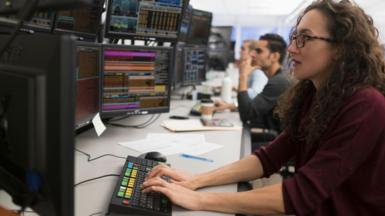 Female trader with long hair and glasses sits at keyboard in front of screens