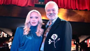 Madonna and Graham Norton are pictured together. She is wearing a blue dress, he is in a dark jacket with a flower motif. They are photographed against a nightclub background.