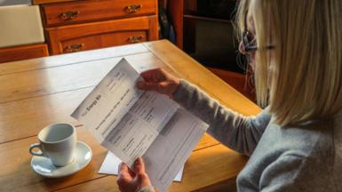 Woman looks at bill at a kitchen table with a cup of tea in front of her.