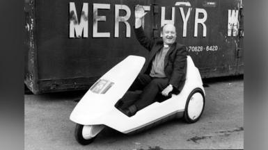 Y Parch Bill Morgan, arweinydd Cyngor Bwrdeistref Merthyr ar y pryd, mewn Sinclair C5 o flaen arwydd Merthyr