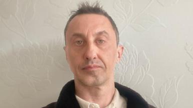 David Robinson is photographed indoors against a pale wall with a subtle floral pattern. He is shown from the shoulders up, wearing a light shirt and a dark jacket. The lighting is soft and even, and the background is uncluttered.