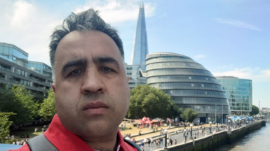 A head shot of a man with short dark hair and red shirt. He is in London with the River Thames, the old City Hall and the Shard in the background. 
