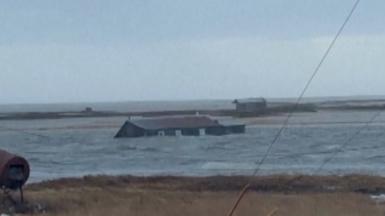 A house submerged almost up to its roof floats in water near land.