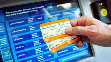 Two kinds of rail tickets are held up in front of a ticket machine displaying a variety of destinations in the UK