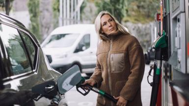 A stock photo of lady in a brown jacket filling up her car with fuel at a petrol station.